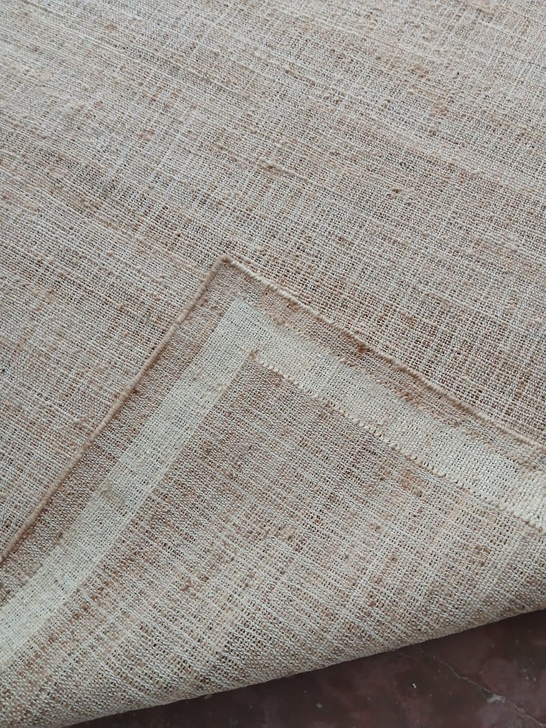 Peut inclure: Gros plan sur un tapis beige tiss&eacute; avec une surface textur&eacute;e. Le tapis a une teinte naturelle et terreuse avec des fibres visibles et un motif subtil. Le coin du tapis est repli&eacute;, r&eacute;v&eacute;lant une bordure plus claire.