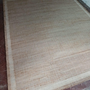 Peut inclure: Un tapis rectangulaire tiss&eacute; de couleur beige naturel. Il pr&eacute;sente une bordure beige plus claire. La texture grossi&egrave;re sugg&egrave;re un mat&eacute;riau durable. Le tapis est pos&eacute; sur un sol brun-rouge. Id&eacute;al pour un salon ou une chambre.