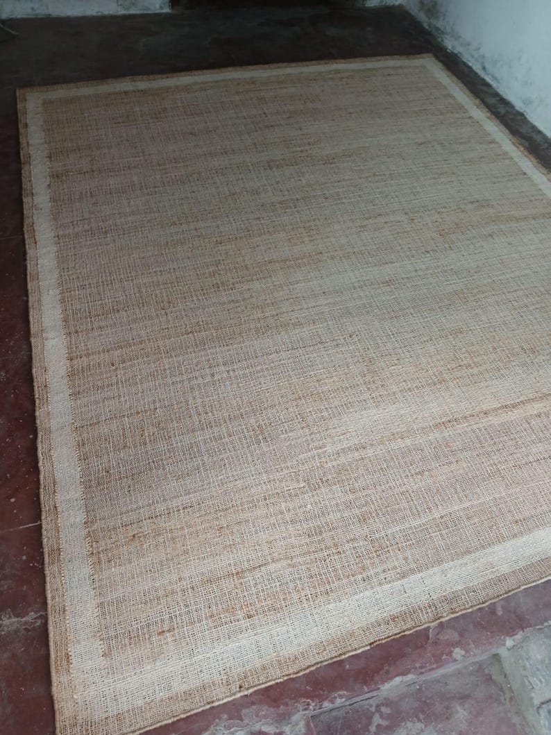 Peut inclure: Un tapis rectangulaire tiss&eacute; de couleur beige naturel. Il pr&eacute;sente une bordure beige plus claire. La texture grossi&egrave;re sugg&egrave;re un mat&eacute;riau durable. Le tapis est pos&eacute; sur un sol brun-rouge. Id&eacute;al pour un salon ou une chambre.