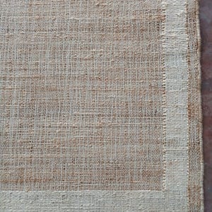 Peut inclure: Gros plan sur un tapis rectangulaire en jute tiss&eacute;, de couleur beige naturel. Le tapis a une bordure beige plus claire. La texture grossi&egrave;re et le tissage visible lui conf&egrave;rent un aspect rustique. Mesures en centim&egrave;tres.