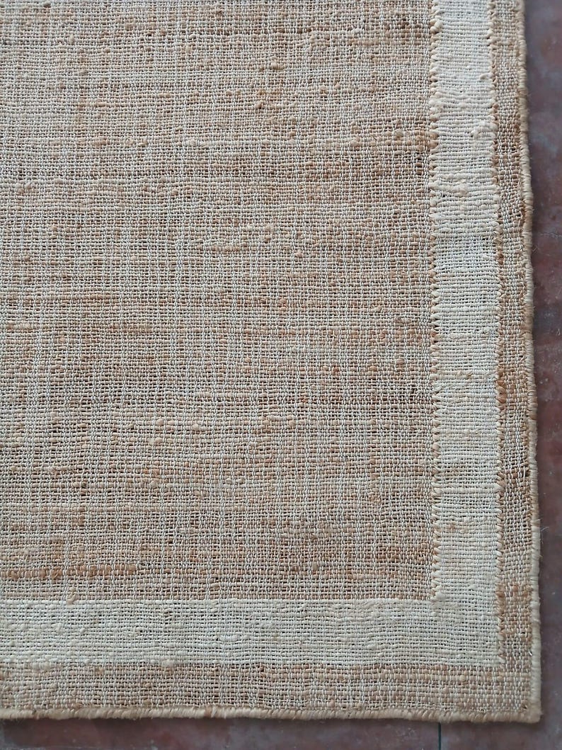 Peut inclure: Gros plan sur un tapis rectangulaire en jute tiss&eacute;, de couleur beige naturel. Le tapis a une bordure beige plus claire. La texture grossi&egrave;re et le tissage visible lui conf&egrave;rent un aspect rustique. Mesures en centim&egrave;tres.