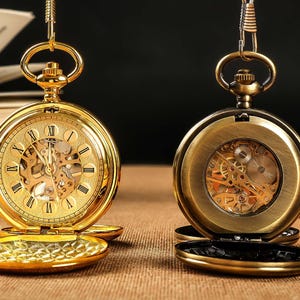Puede incluir: Dos relojes de bolsillo dorados con esferas abiertas, que muestran intrincados movimientos mecánicos y números romanos. Un reloj está completamente abierto, revelando el mecanismo interno, mientras que el otro está parcialmente abierto. Ambos están suspendidos por cadenas.