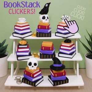 May include: A display of miniature book stacks with various toppers, including a crow, skull, and witch's hat. The stacks are made of colorful books and are arranged on a white shelf. The text "BookStack CLICKERS!" is at the top.