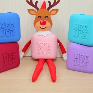 May include: A plush reindeer doll with a red nose and white collar, wearing a pink square Nee Doh block as a body. It is surrounded by four additional square Nee Doh blocks in blue, red, teal, and purple, each embossed with the text Nee Doh.