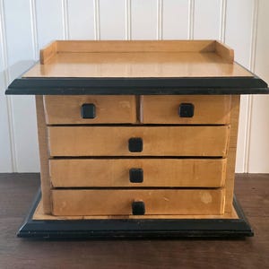 May include: A wooden desktop organizer featuring a light natural wood finish with contrasting black trim and black square knobs. The unit includes two small top drawers, three wider drawers below, and a flat top surface with a raised back edge.