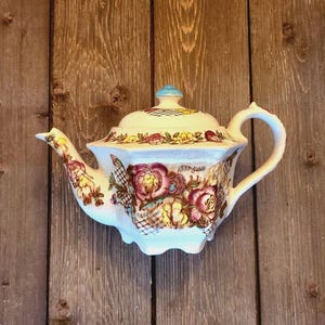 May include: A white teapot with a floral pattern in shades of pink, yellow, and brown. The teapot has a blue knob on the lid and a scalloped edge.