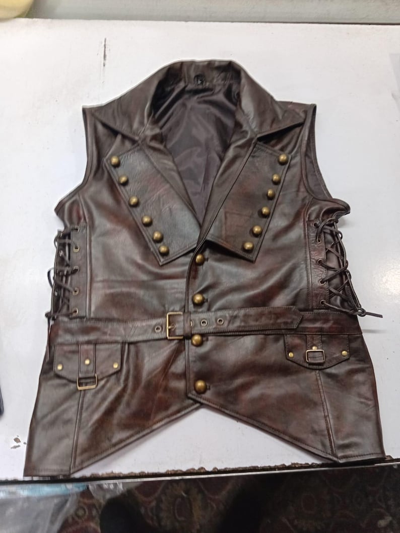 May include: A brown leather vest with a notched collar and brass-colored buttons. The vest has a belt with a buckle and two pockets. The sides of the vest have a lace-up design. This vest is a steampunk-inspired garment.