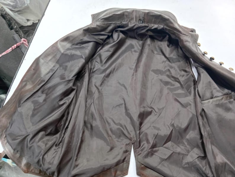 May include: A brown leather jacket with a dark brown satin lining. The jacket is open, revealing the interior lining and the button details along the front. The jacket appears to be vintage or pre-owned.