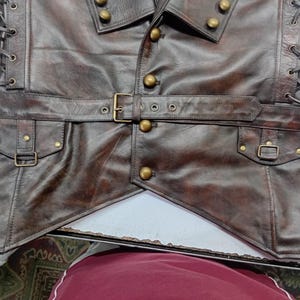 May include: A brown leather vest with a steampunk design. The vest features a notched collar, brass buttons, a belt with a buckle, and pockets with small buckles. The sides are laced up with leather cord.
