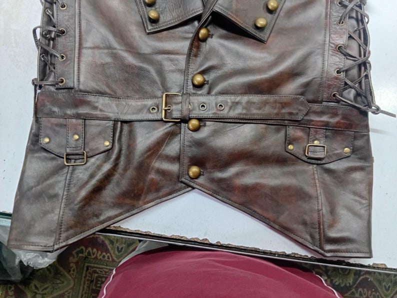 May include: A brown leather vest with a steampunk design. The vest features a notched collar, brass buttons, a belt with a buckle, and pockets with small buckles. The sides are laced up with leather cord.