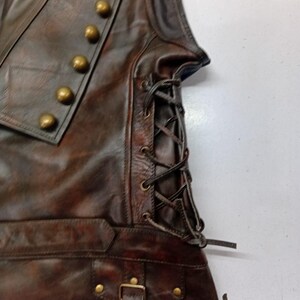 May include: A brown leather vest with brass-colored buttons and a lace-up side. The vest has a pocket with a buckle closure and is made of distressed leather.