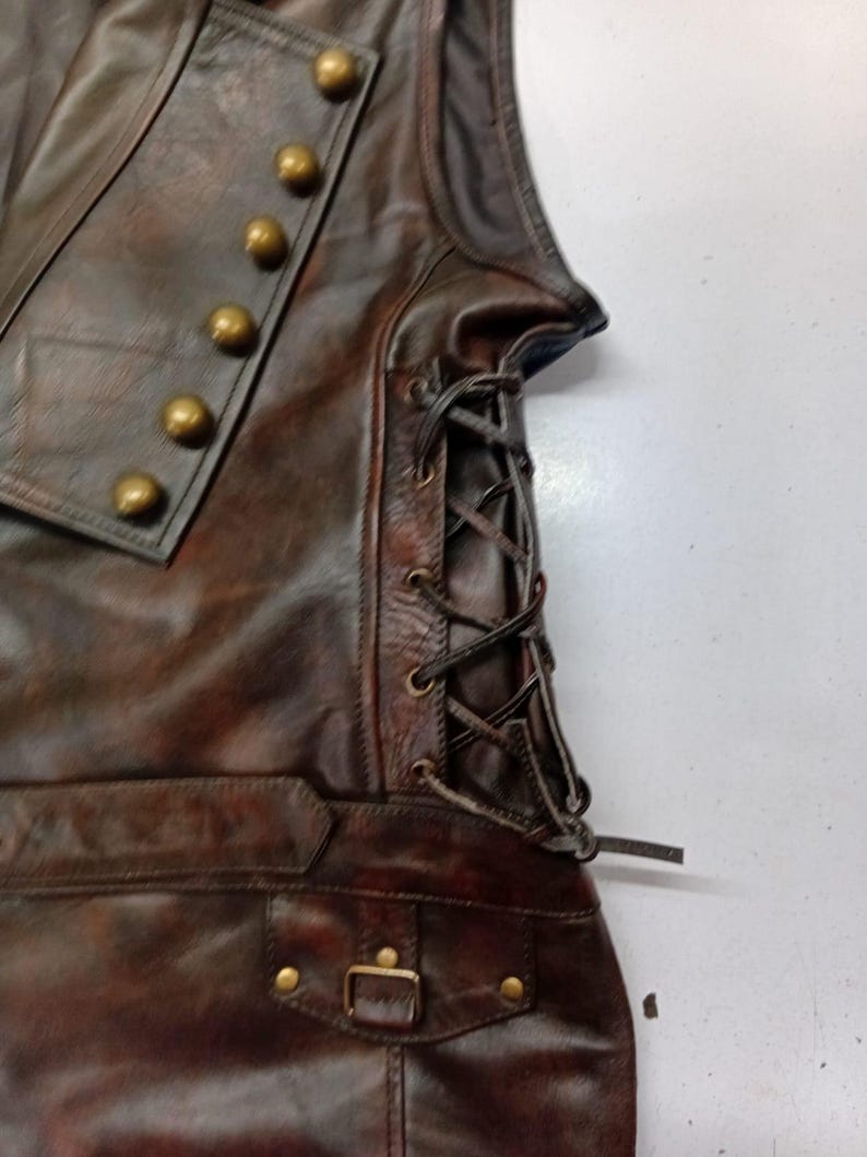 May include: A brown leather vest with brass-colored buttons and a lace-up side. The vest has a pocket with a buckle closure and is made of distressed leather.