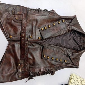May include: A brown leather vest with a steampunk aesthetic. The vest features multiple pockets, brass-colored buttons, and adjustable straps with buckles. The vest has a distressed finish and a dark interior lining.