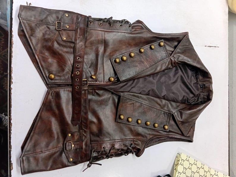 May include: A brown leather vest with a steampunk aesthetic. The vest features multiple pockets, brass-colored buttons, and adjustable straps with buckles. The vest has a distressed finish and a dark interior lining.