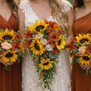 以下が含まれることがあります： ひまわり、バラ、緑の葉のブーケを持つ人々のグループ。ブーケには、黄色のひまわり、オレンジ色のバラ、その他の花が含まれています。人々は錆色のドレスを着ています。ブーケはおそらく結婚式用です。