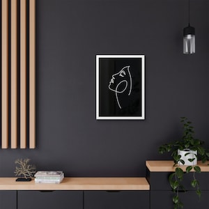 Könnte beinhalten: Ein gerahmter Kunstdruck mit einer minimalistischen Strichzeichnung eines Frauenprofils in Weiß auf schwarzem Hintergrund. Das Kunstwerk ist in einem weißen Rahmen eingefasst, was einen auffälligen Kontrast erzeugt. Das Stück wird an einer dunklen Wand ausgestellt.