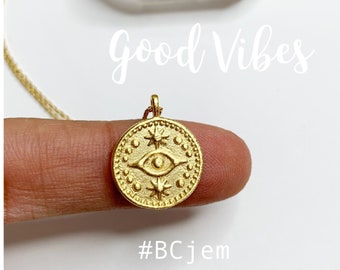 May include: Gold necklace with a round pendant featuring a stylized eye with stars and dots. The pendant is on a finger and the text "Good Vibes" and "#BCJem" are below it.