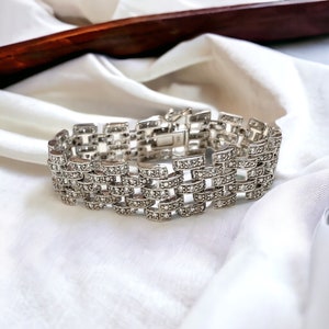Pode incluir: Um bracelete de prata com um design de corrente de elos, com pequenas pedras transparentes engastadas no metal. O bracelete está sobre um fundo de tecido branco.