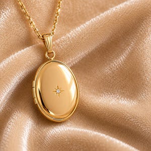 May include: A polished gold oval locket pendant suspended from a matching gold chain. The locket features a smooth, reflective surface with a small, centred starburst engraving containing a single, tiny embedded clear gemstone at its core.