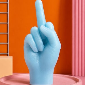 Può includere: Una candela in cera azzurra a forma di mano che fa il gesto del dito medio. La candela presenta una superficie liscia di colore uniforme con un unico stoppino bianco che sporge dalla punta del dito medio esteso, posizionata su una base.