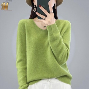 May include: A light green V-neck sweater with long sleeves. The sweater has a soft, textured appearance and a ribbed cuff and hem. The model is wearing white trousers. The sweater is a solid colour and appears to be made of a warm material.