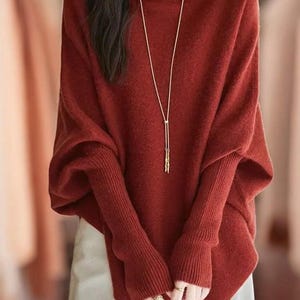 Könnte beinhalten: Ein rostfarbener Pullover mit einer langen goldenen Halskette. Der Pullover hat lange Ärmel mit gerippten Bündchen und einen weiten, drapierten Körper. Das Model trägt eine weiße Hose. Der Pullover ist einfarbig und scheint aus einem weichen Material zu sein.