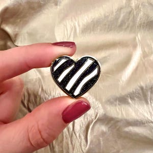 May include: A heart-shaped pin with a black and white striped design. The pin has a gold-colored border. The heart is held up against a gold-colored, textured background.