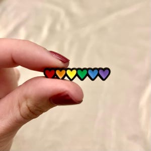 May include: A small, black accessory featuring a row of seven colorful hearts in a rainbow spectrum. The hearts are arranged horizontally, with each heart a different color: red, orange, yellow, green, blue, and purple. The accessory is held between fingers.
