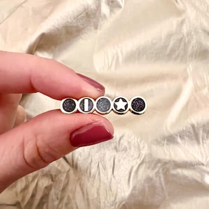 Könnte beinhalten: Goldfarbene Haarnadel mit fünf kreisförmigen Designs. Die Designs umfassen ein dunkles, gesprenkeltes Muster, eine weiße vertikale Linie, ein dunkles, gesprenkeltes Muster, einen weißen Stern und ein dunkles, gesprenkeltes Muster. Die Spange wird von einer Hand mit bordeauxrotem Nagellack gehalten.