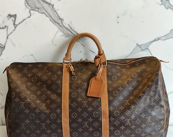 May include: A brown travel bag featuring a repeating monogram pattern in dark brown and tan. The bag includes tan leather handles, vertical leather trim, a gold-tone metal padlock attached to the zipper, and a matching tan leather luggage tag.