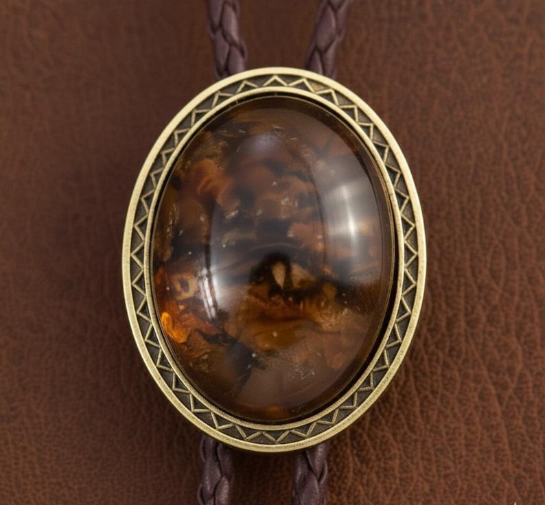 May include: A bolo tie featuring an oval-shaped, polished amber-colored stone set within a decorative antique brass frame with a geometric border. The pendant is attached to a dark brown braided leather cord.