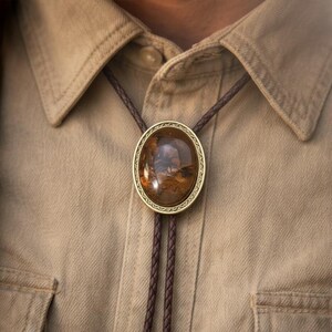 May include: A bolo tie with a large, oval-shaped, polished stone pendant. The stone is a mix of brown and amber hues, set in a gold-colored metal frame. The braided cord is a dark brown color, and the tie is worn on a tan button-down shirt.
