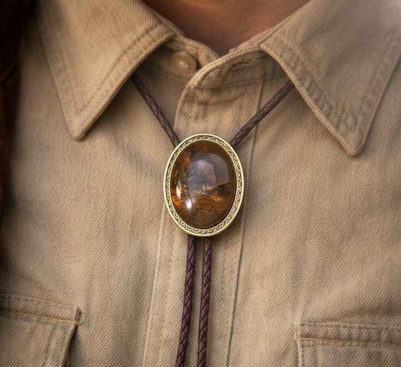 May include: A bolo tie with a large, oval-shaped, polished stone pendant. The stone is a mix of brown and amber hues, set in a gold-colored metal frame. The braided cord is a dark brown color, and the tie is worn on a tan button-down shirt.