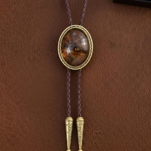 May include: A bolo tie with a brown braided leather cord and a gold-colored oval pendant. The pendant features a brown and amber-colored stone with a decorative gold border. The tips of the cord are also gold-colored and have an engraved design.