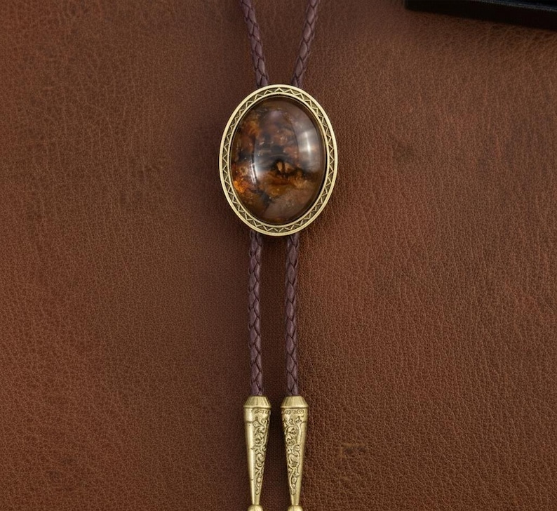 May include: A bolo tie with a brown braided leather cord and a gold-colored oval pendant. The pendant features a brown and amber-colored stone with a decorative gold border. The tips of the cord are also gold-colored and have an engraved design.