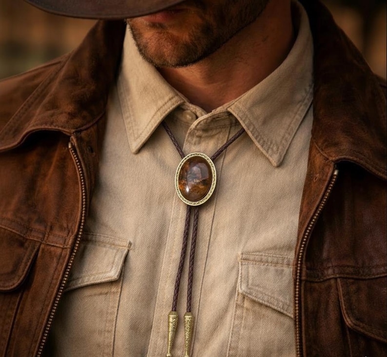 May include: A brown bolo tie with an oval-shaped, patterned stone pendant set in a gold-colored frame. The tie features a braided leather cord and decorative gold-colored tips. The bolo tie is worn over a tan button-down shirt and a brown leather jacket.