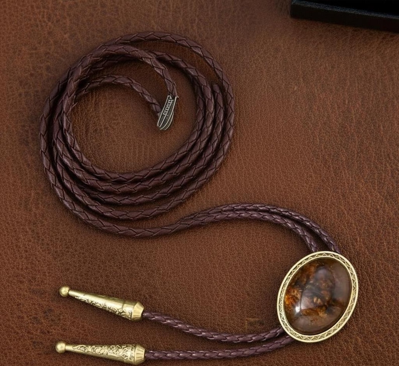 May include: A brown bolo tie with a braided leather cord. The tie features a gold-toned oval pendant with a brown and gold stone. The cord ends have decorative gold-toned tips.