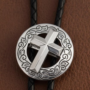 May include: A silver bolo tie featuring a cross design. The cross is centered within a decorative circular frame with an ornate, swirling pattern. The tie has a black braided cord.
