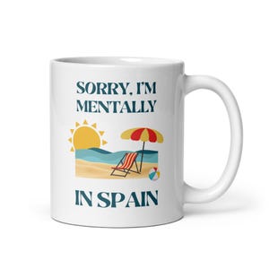 Może przedstawiać: Biały ceramiczny kubek z napisem "SORRY, I'M MENTALLY IN SPAIN" w kolorze granatowym. Kubek przedstawia scenę plażową ze słońcem, falami, leżakiem, parasolem i piłką plażową.