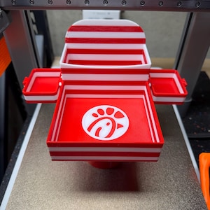May include: A 3D printed multi-tiered tray featuring horizontal red and white stripes. The central square base displays a white Chick-fil-A logo on a red background. Two smaller rectangular side trays are attached to the main structure.