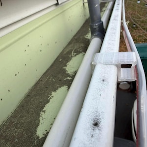 May include: A clear plastic gutter clip is attached to a white gutter. The gutter is mounted on a light green surface. A white pipe runs parallel to the gutter. The background includes a green trash can and trees.