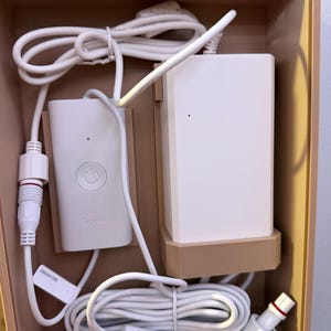 May include: A white Govee control unit and power adapter housed in a tan plastic tray. The set includes a rectangular controller with a power button, a larger rectangular power block, and white connecting cables with threaded screw-on connectors.
