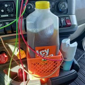 May include: A plastic jug of Icy Tea with a yellow cap sits inside a small, orange, square-shaped plastic crate holder. The jug features an orange label with the text Original Icy Tea. The crate is positioned on a dark gray vehicle dashboard.