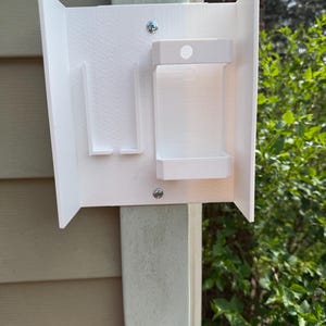 Könnte beinhalten: Ein weißer, an der Wand montierter Spender mit rechteckigem Körper und kleinem Regal. Der Spender ist mit Schrauben an einem vertikalen Pfosten befestigt. Das Design umfasst zwei vertikale Schlitze und eine kleine, kreisförmige Öffnung oben.