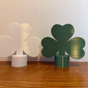 Puede incluir: Dos decoraciones de trébol, una blanca y otra verde, cada una con tres hojas en forma de corazón. Los tréboles están montados sobre bases cilíndricas. Los objetos se exhiben sobre una superficie de madera.