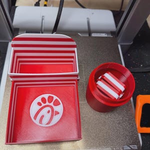 May include: A 3D printed red and white striped box with a hinged lid and a white chicken logo on the interior base, alongside a separate red cylindrical container holding smaller red and white striped components on a textured print bed.