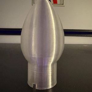 Op de afbeelding: Een licht zilverkleurig, druppelvormig object met een cilindrische basis. Het object heeft een geribbelde textuur en een kleine inkeping in de basis. Het is waarschijnlijk een decoratief item of een onderdeel van een groter product.