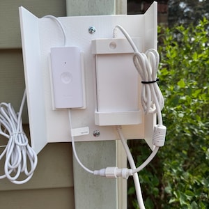 May include: White electronic device mounted on a white bracket, attached to a beige exterior wall. The device includes a power button and a rectangular component, connected by white cords. The background features green foliage.