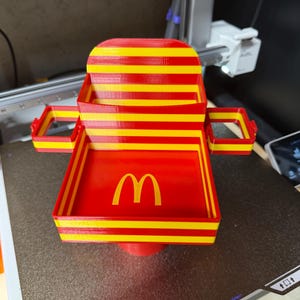May include: A 3D-printed red and yellow striped plastic cup holder and tray accessory featuring the yellow McDonald's golden arches logo on the base. The object has a central square tray, a vertical back panel, and two side-mounted square cup holders.