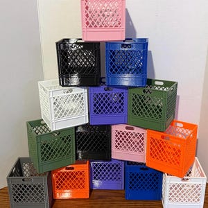 May include: A collection of small, colorful plastic crates in various shades, including pink, black, blue, white, purple, green, orange, and gray. The crates are stacked, showcasing their diamond-patterned sides and handle openings, suitable for storage or display.